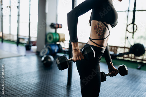 Slender body of a young woman with tattoo in a black sportswear that is doing exercises with dumbbells in the gym
