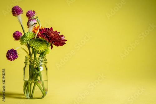 Fototapeta Naklejka Na Ścianę i Meble -  Small glass jar with clover and mums on a yellow background.