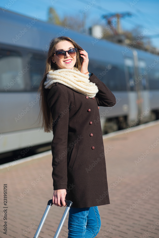 Fototapeta premium Happy young woman wearing glasses dressed in coat with big travel bag at the railway station on the train background