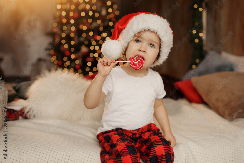 Obraz premium Little cute baby in Santa Claus hat is sitting in the room on the background of the Christmas tree and eating sweet candy on a stick
