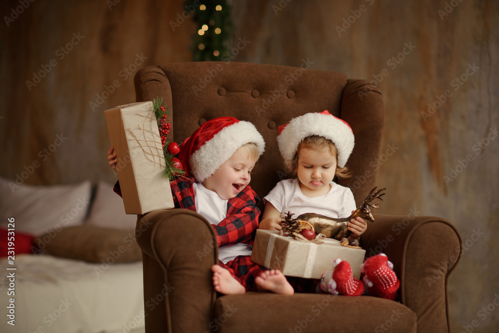 Little cute kids, brother and sister, wearing Santa Claus hats, are sitting on a big easy chair in the living room and are looking at Christmas presents in boxes. 