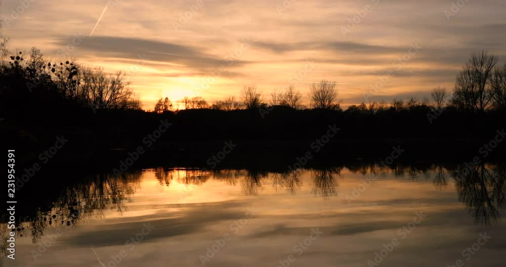 The orange sunset over the lake in the dark colors and the strong shadows of the sun on the horizon fits behind the trees and the sky is full of color clouds. The orange sunset over the lake in the dark colors and the strong shadows of the sun on the horizon fits behind the trees and the sky is full of color clouds.