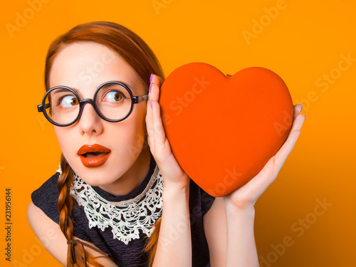 surprised young girl in glasses with two pigtails holding heart shape box on yellow background