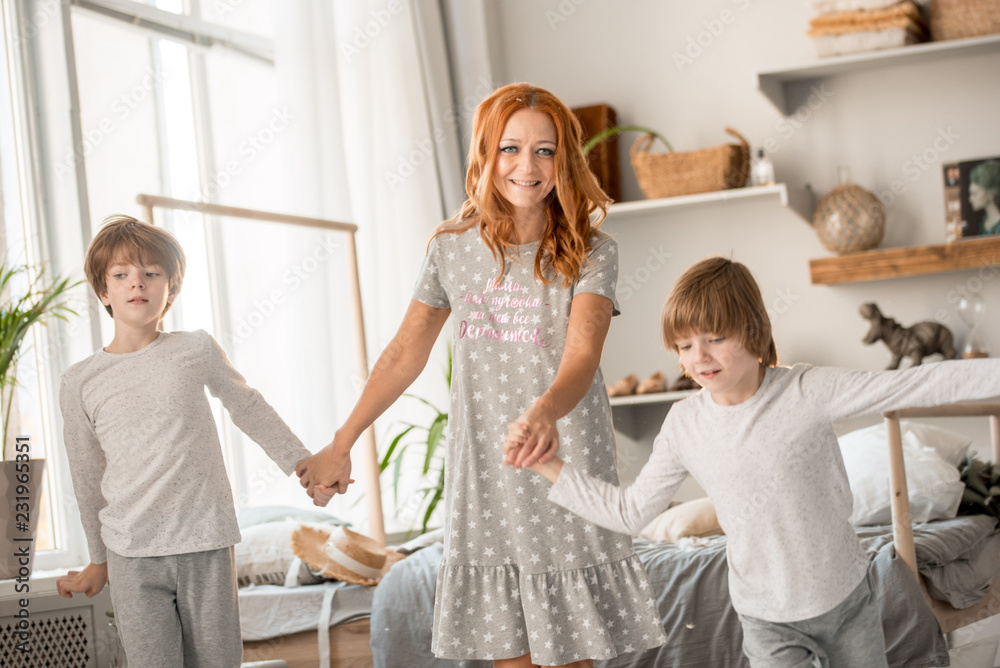 Fototapeta premium blonde caucasian baby two years old in brunette woman mother arms breastfeeding sitting on bed white clothes in bedroom next to window indoor. Woman is back.