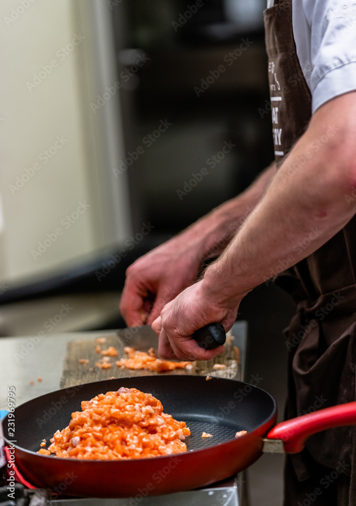 Red Pan with Pile of Cutted Salmon Pieces in the Foreground, Male Chef Cutting Fresh Salmon on the Wooden Board in the Background Red Pan with Pile of Cutted Salmon Pieces in the Foreground, Male Chef Cutting Fresh Salmon on the Wooden Board in the Background