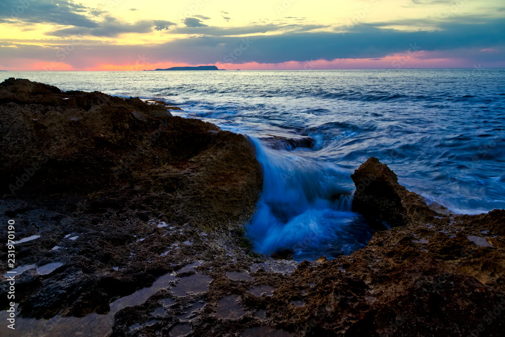 sunset over the sea, pink clouds, rocks in the foreground, which is flooded with a wave