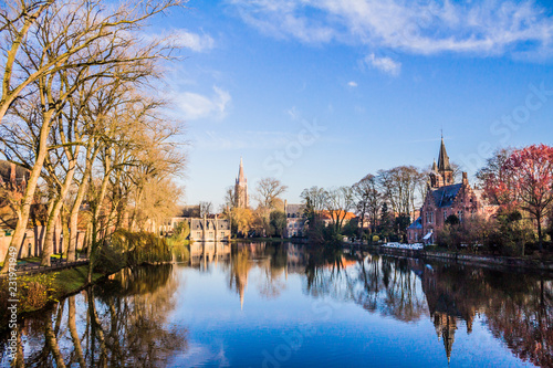 Cityscape in a part of Bruges, Minnewater lake park surrounded by baretrees, buildings and medieval castle, mirror reflection on water surface, sunny winter day in province of West-Vlaanderen, Belgium