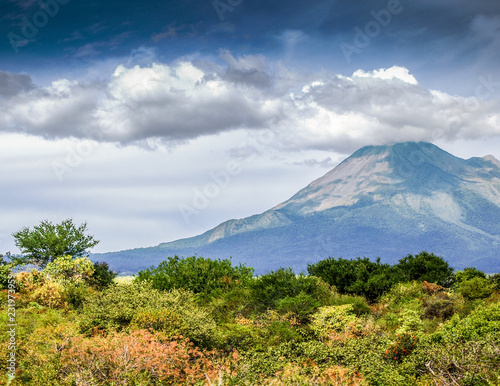 Mexican landscape with a hill with green vegetation and the Colima volcano in the background  sunny day with a blue sky and abundant clouds in the state of Jalisco Mexico