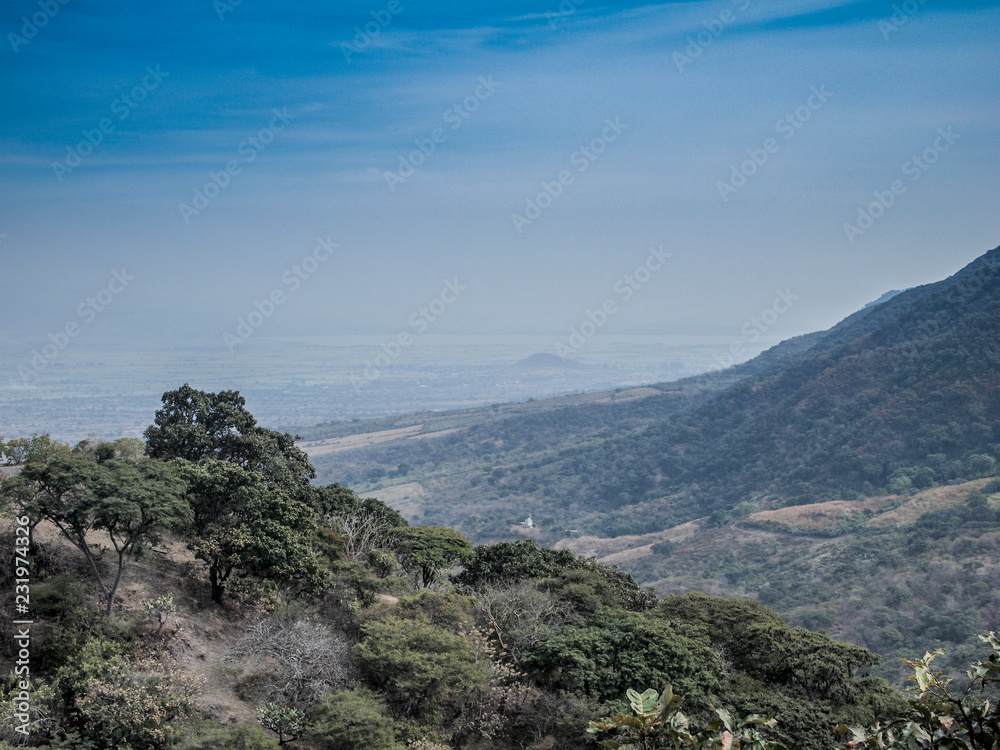 Scenic aerial view of Sierra Madre Occidental against hazy blue sky, green vegetation and trees, dry summer day at Volcanic Belt mountain range system between Jalisco and Colima states, Mexico