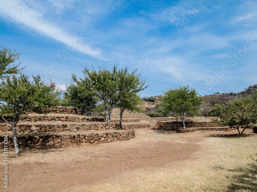 Small circular pyramids, green trees against blue sky, Guachimontones pre-Hispanic archaeological zone, stepped conical structures, mountain in background, day in arid land, Teuchitlan Jalisco Mexico photo