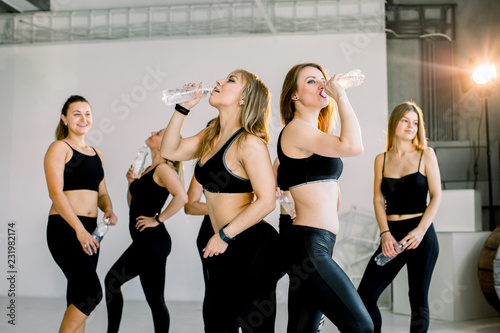 Group of young sporty girls with water boottles standing at white wall. Female companions in gym resting after fitness, indoor full length, loft studio, copy space © sofiko14