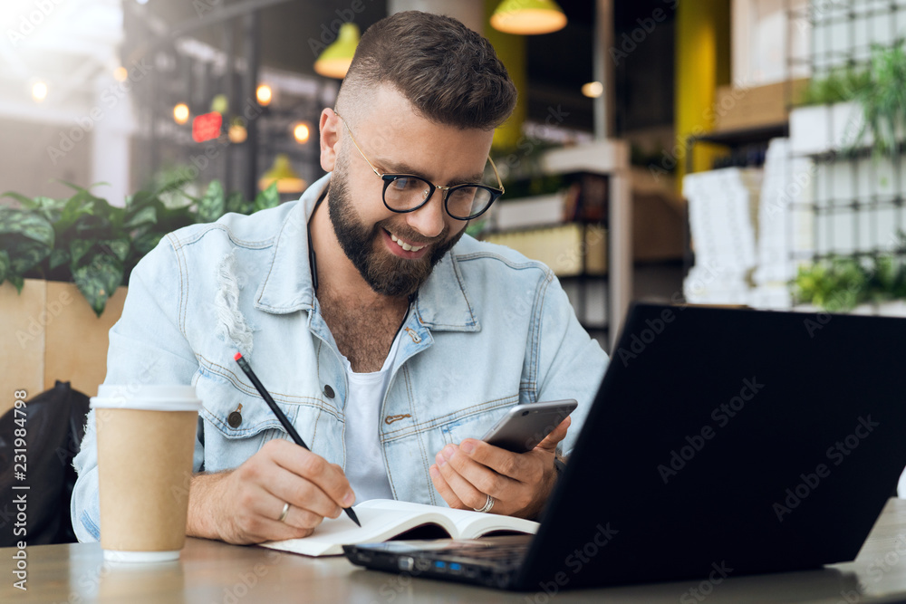 Bearded hipster man sits at table in front of laptop and writes in notebook,student prepares for exams,freelancer works. Bearded hipster man sits at table in front of laptop and writes in notebook,student prepares for exams,freelancer works.