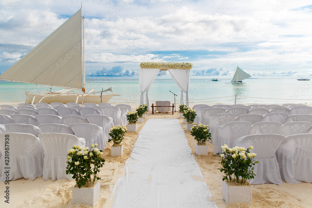 Wedding ceremony on a tropical beach in white. The arch is decorated with flowers on the sandy beach, decorated chairs for guests, sand castle and yacht in the sea. Wedding and honeymoon concept.
