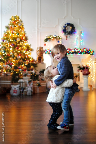 Дети ждут чудо в Новый год. С Новым годом! Счастливого Рождества! Елка с новогодними игрушками. Маленький мальчик обнимает девочку возле елки с игрушками. Брат с сестрой.