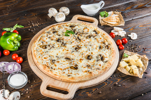 Large creamy pizza with mushrooms and chicken on a round cutting board on a dark wooden background. Pizza ingredients