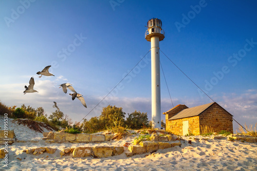 Landscape with sea lighthouse on the coast during sunrise near the ruins on sandy beach photo