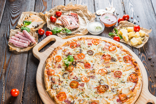 Large meat pizza with bacon, ham, chicken, sausage and tomatoes on a round cutting board on a dark wooden background. Food Ingredients.