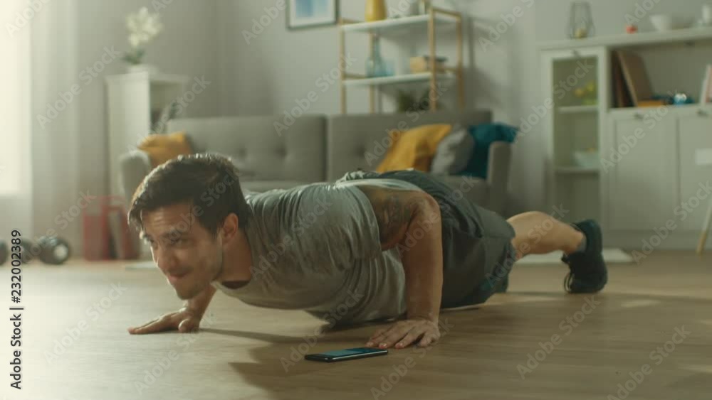 Athletic Fit Man in T-shirt and Shorts is Doing Push Up Exercises While Using a Stopwatch on His Phone. He is Training at Home in His Living Room with Minimalistic Interior.