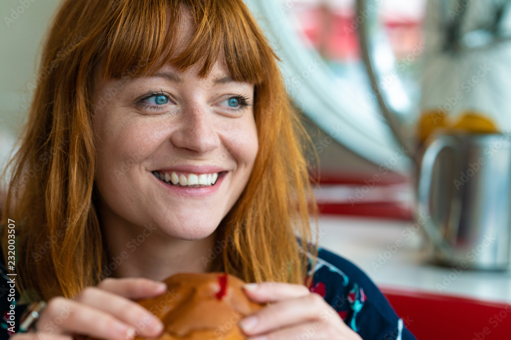 Portrait of ginger girl with blue eyes on a floral dress in diner eating a burger and smiling Portrait of ginger girl with blue eyes on a floral dress in diner eating a burger and smiling