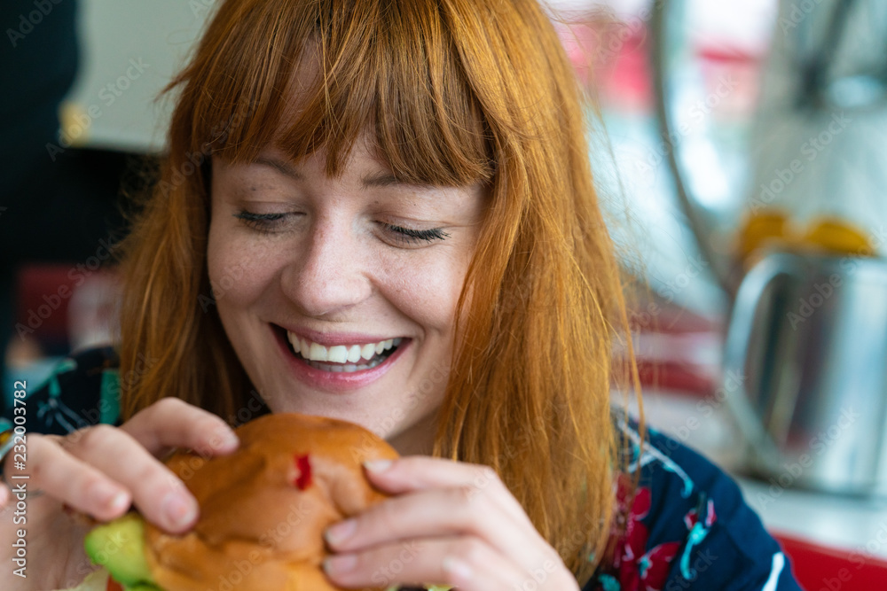 Portrait of ginger girl with blue eyes on a floral dress in diner eating a burger and smiling Portrait of ginger girl with blue eyes on a floral dress in diner eating a burger and smiling