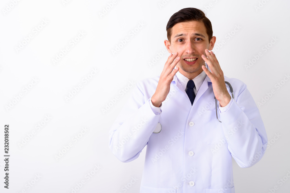Studio shot of young handsome man doctor looking shocked against Studio shot of young handsome man doctor looking shocked against