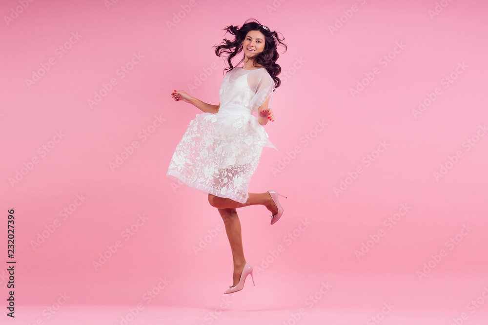 young woman with curly hair wearing white dress on a pink background in the studio. girl jumping for joy young woman with curly hair wearing white dress on a pink background in the studio. girl jumping for joy