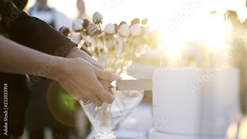 Groom and bride cuting first place of cake during wedding event. Close-up hands together holding knife and making first cut on white wedding cake in sun shining