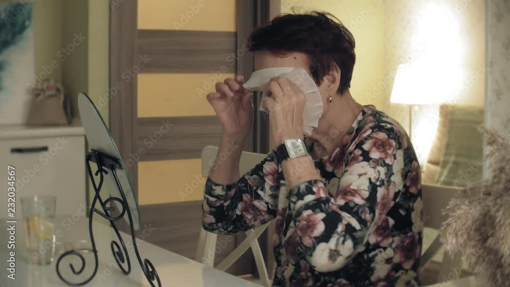 An old woman looks in a mirror, removes a cosmetic tissue mask from her face. Facial care. Plastic surgery and collagen injections. Realistic images with their own imperfections.