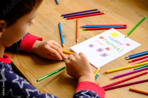 A boy sits at a wooden table on which is a Mother's Day postcard and colored pencils.