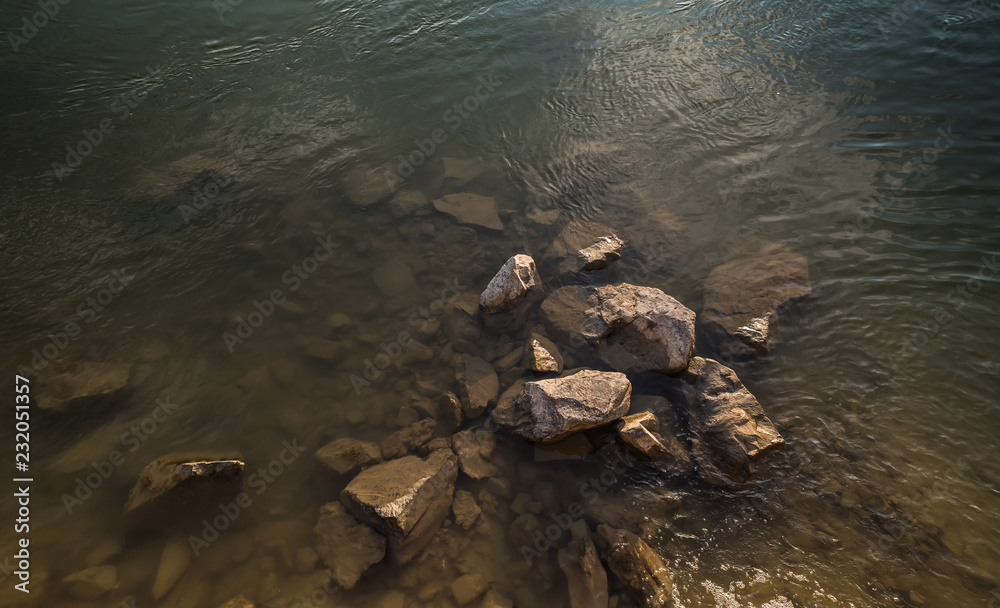 Top view of a cluster of big, aged, worn stones covered with algae, clearly seen in shallow river water, lit with warm and soft sunset light.