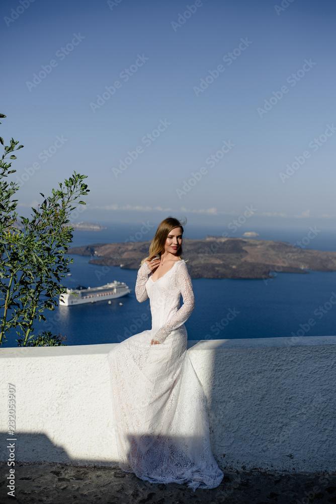 beautiful slim blond girl woman top model bride with long hair and legs in the white bridal ball gown with a long train and flounces stands on a picturesque island of Santorini in Greece