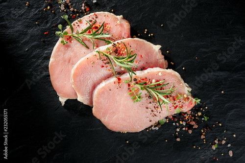 Slices of raw pork loin with the addition of aromatic herbs and spices on a black stone background, top view