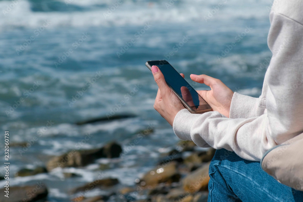 Woman is typing a message on phone sitting on the sea shore in autumn. Hands close-up.