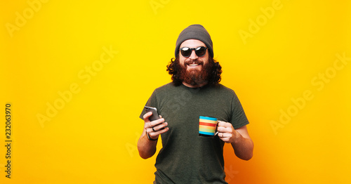 Happy bearded hipster man using phone and holding cup of coffee