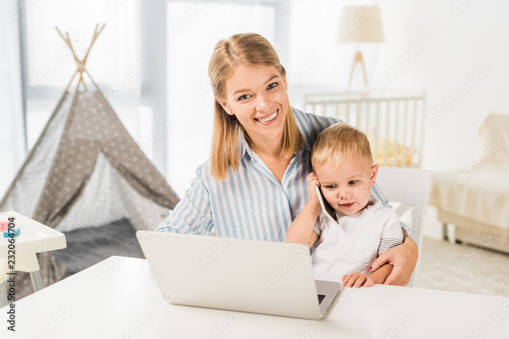 cute toddler holding smaptphone with mother sitting at desk while using laptop cute toddler holding smaptphone with mother sitting at desk while using laptop