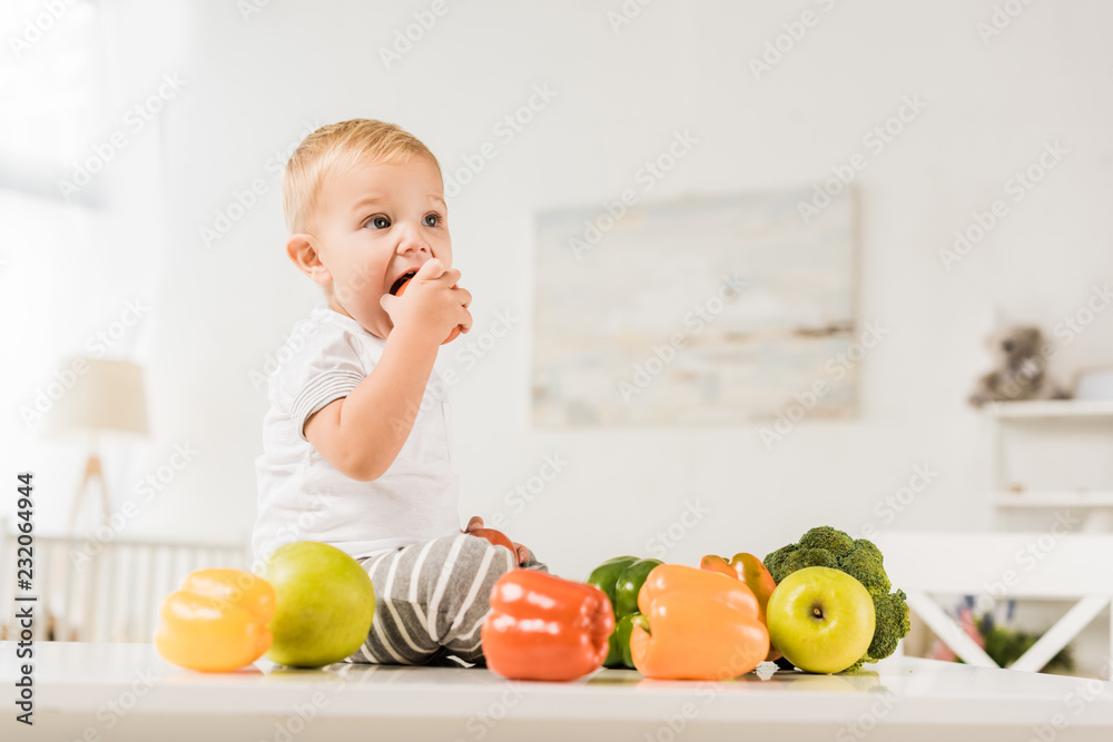 Fototapeta premium cute toddler eating and sitting on table surrounded by fruit and vegetables