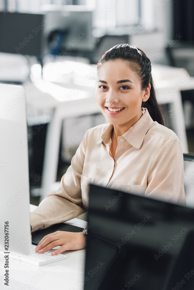beautiful young businesswoman using desktop computer and smiling at camera in office