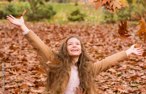 A teenager girl in a coat laughs and throws up leaves in autumn park against the background of yellow foliage.