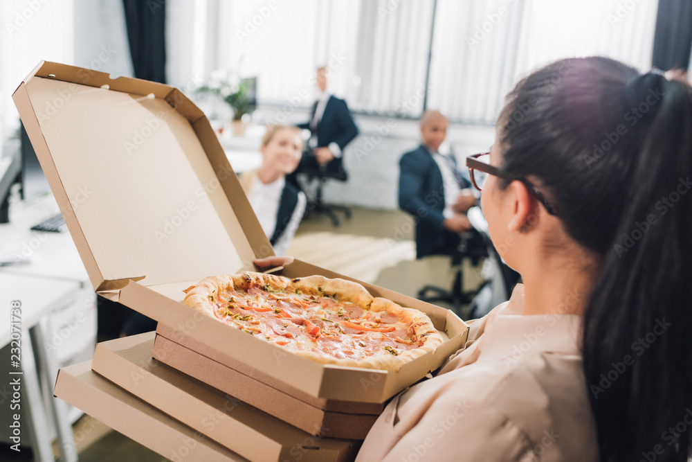 young woman holding pizza boxes and looking at colleagues in open space office