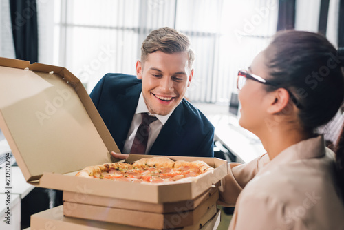 young businesswoman holding pizza boxes and smiling businessman looking at tasty pizza