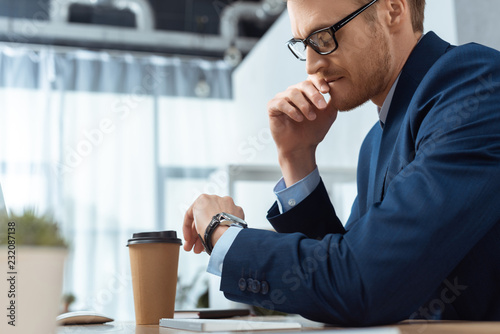 Photography serious businessman in eyeglasses checking watch at table with paper coffee cup