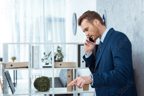serious businessman in suit talking on smartphone and gesturing by hand in modern office