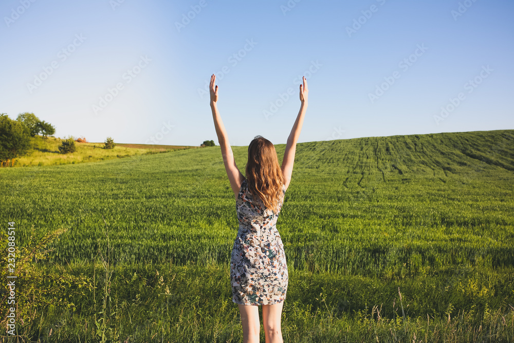 Beautiful girl is doing yoga on summer green grass field. Happy woman in rustic dress puts her hands up in asana. Freedom and harmony concept.