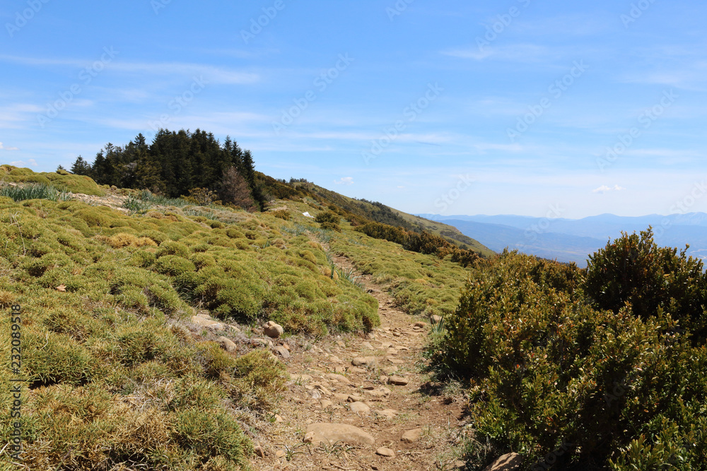 A landscape of a path on the edge of Peña Oroel mount, with the Pyrenees as background, a wide valley with blue sky and some bushes, in Aragon, Spain