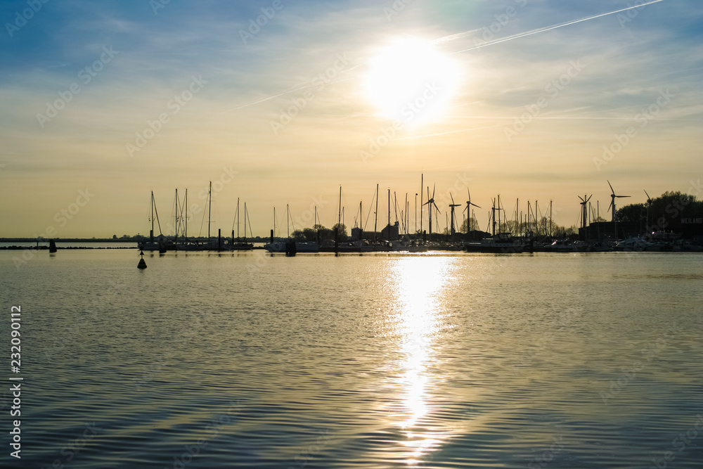 Lovely view of the sun setting behind the harbour leaving a silhouette of wind turbines and sailing boats with their mast on Fehmarn island in Germany. The sunlight is shimmering on the water surface.