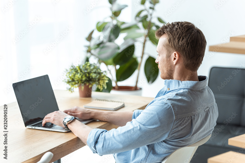 selective focus of young businessman working on laptop with blank screen at table in office selective focus of young businessman working on laptop with blank screen at table in office