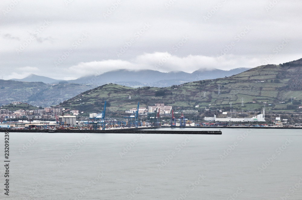 Zierbena port with green hills as background, shot from Paseo de la Galea promenade in Getxo, Basque Country, Spain, during a foggy cloudy winter day