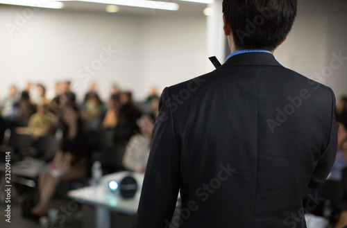 "Speaker on Stage Presenting at Event. Audience at Conference Hall Lecture Series. Corporate Presenter Presentation on Healthcare Initiatives. Crowd of People Blurred De-focused. Expert Public Speech
