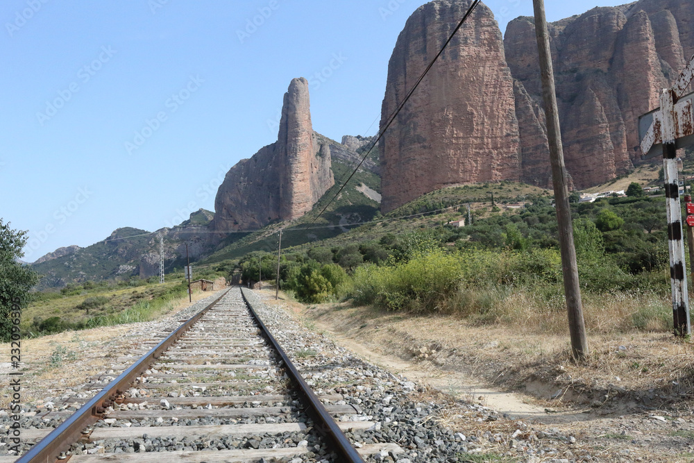 A railway and a light pole during a sunny summer, with the high Mallos de Riglos rock formations and mountains on the background, in Aragon, Spain