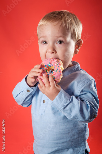 close up of one young blond and cute baby eating and holding with his hands one pink chocolate doughnut over a red background in studio